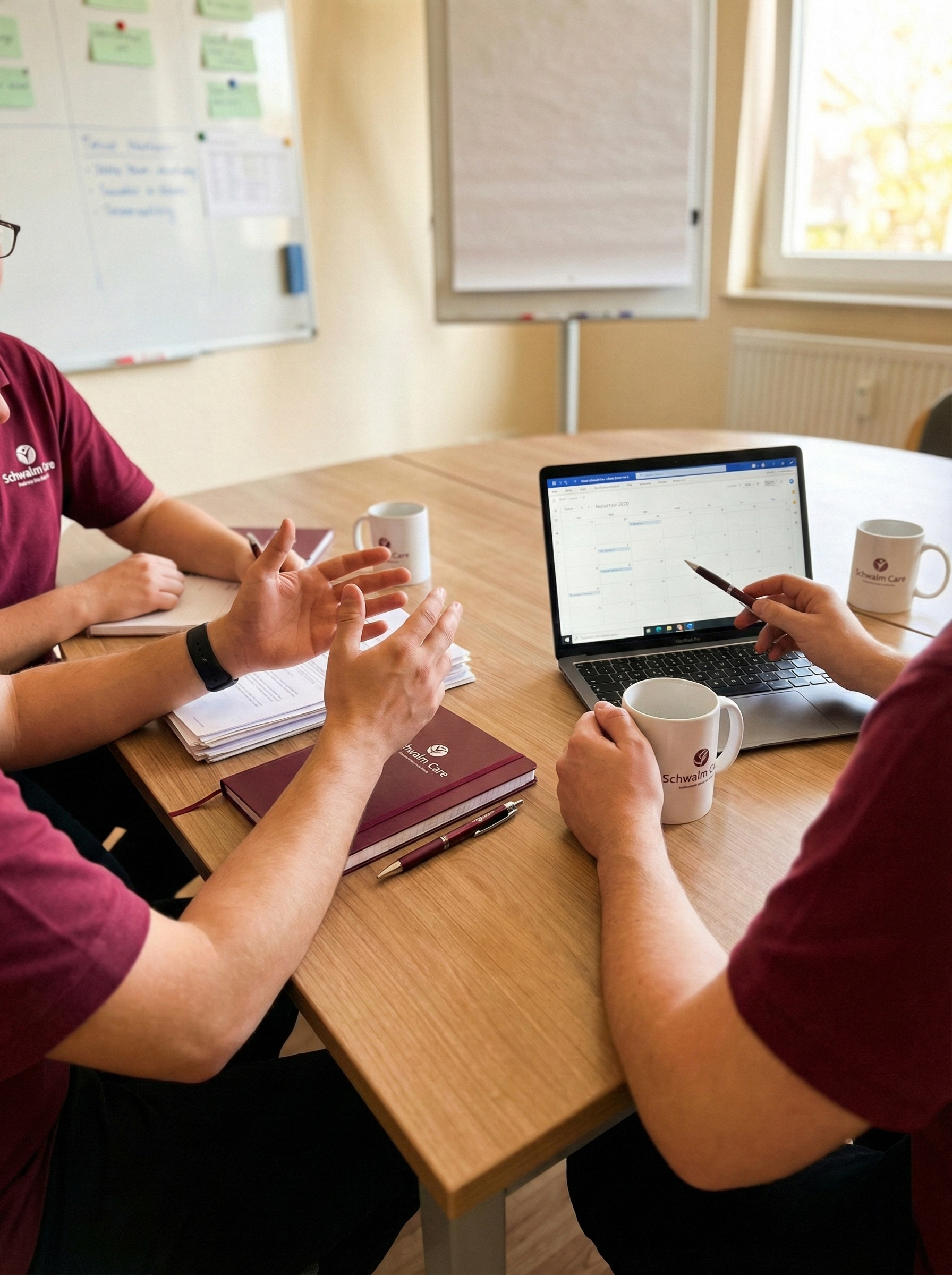 Nahaufnahme einer Teambesprechung am Holztisch: Drei Personen in weinroten Schwalm Care Polo-Shirts sind zu sehen, deren Gesichter nicht erkennbar sind. Eine Person gestikuliert mit den Händen, eine andere zeigt mit einem Stift auf einen Laptop-Bildschirm, der einen Kalender anzeigt. Auf dem Tisch liegen Notizbücher, Stifte und weiße Tassen mit dem Schwalm Care Logo. Im Hintergrund sind unscharf Whiteboards und ein Fenster zu sehen.