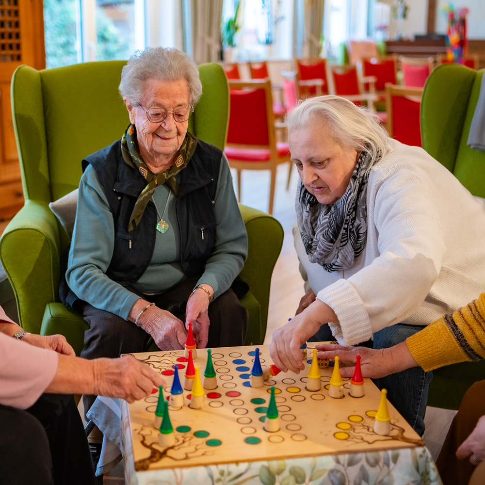 Schwalm Care - Tagespflege - Mehrere ältere Menschen sitzen zusammen und spielen ein Brettspiel an einem Tisch in einem Gemeinschaftsraum